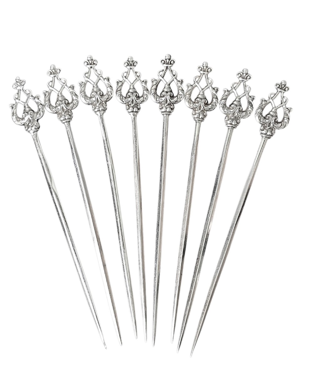A set of eight sterling silver hors doeuvre skewers arranged in a fan shape, featuring ornate openwork scrollwork handles and tapered pins.