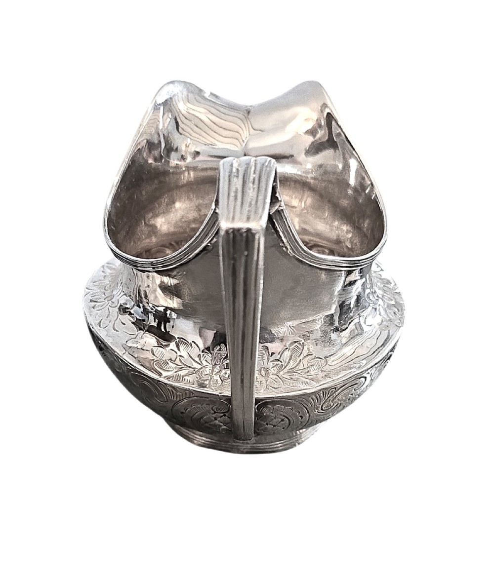 A George III sterling silver cream jug with chased scroll decoration, an angular high loop handle, and a circular foot. View II