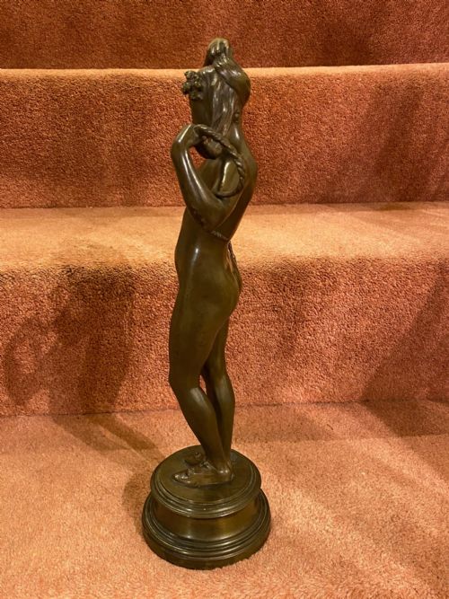 fig 1. A dark brown bronze statuette of a standing female nude holding her long plaited braids, mounted on a moulded circular pedestal base.