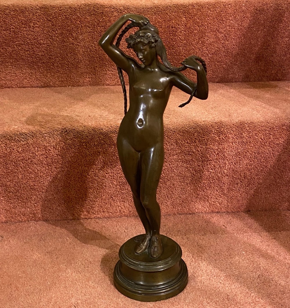 A dark brown bronze statuette of a standing female nude holding her long plaited braids, mounted on a moulded circular pedestal base.