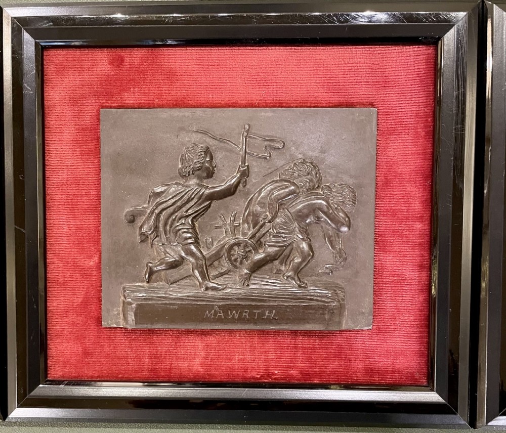 A set of framed hand-carved dark Welsh slate relief plaques mounted against red fabric backgrounds within black moulded frames. View V