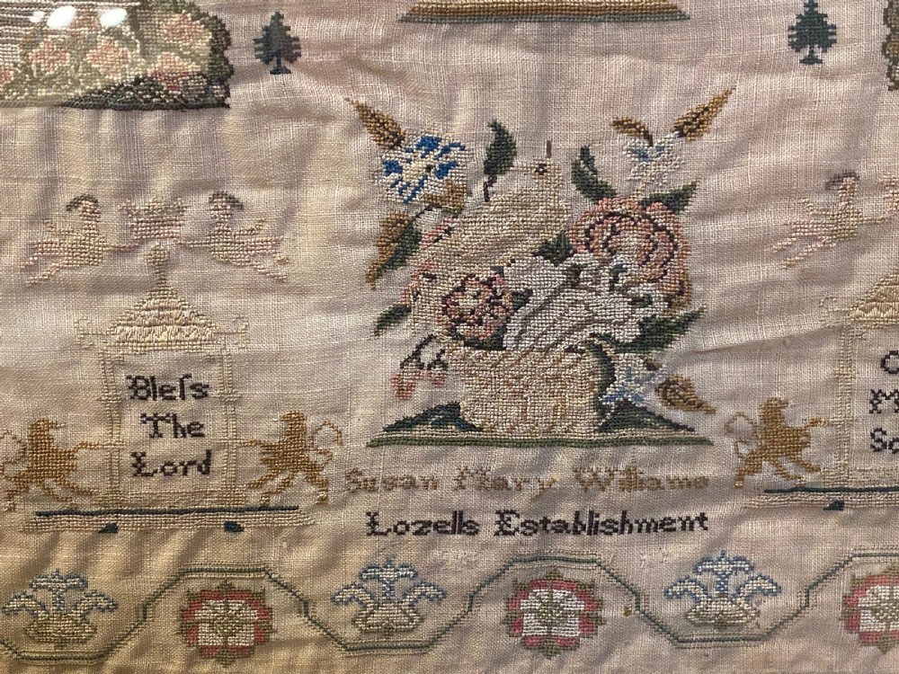 A framed Georgian needlework sampler featuring embroidered religious verses, floral borders, stylized trees, and animal motifs on a beige woven background. View III