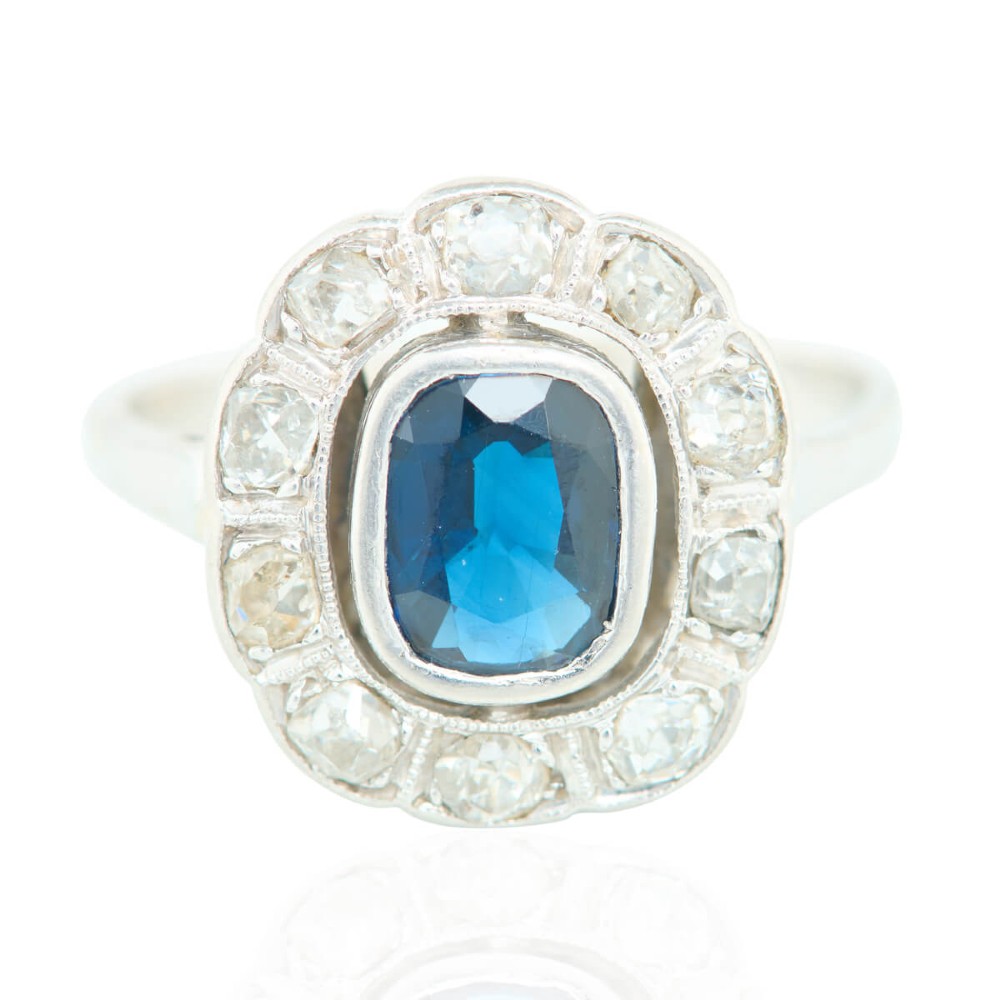 An 18ct white gold cluster ring featuring a central cushion cut blue sapphire framed by a halo of old cut diamonds with millgrain detailing.