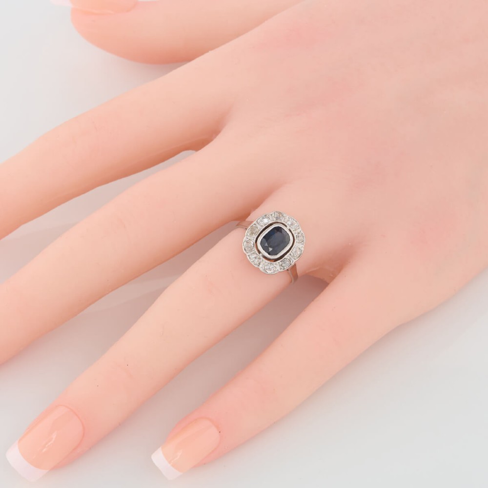 An 18ct white gold cluster ring featuring a central cushion cut blue sapphire framed by a halo of old cut diamonds with millgrain detailing. View II