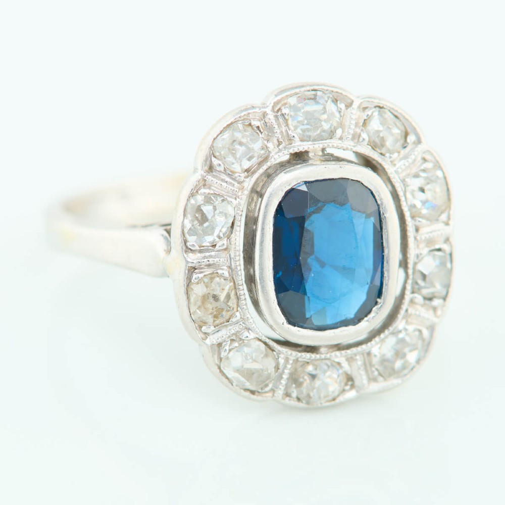 An 18ct white gold cluster ring featuring a central cushion cut blue sapphire framed by a halo of old cut diamonds with millgrain detailing. View III