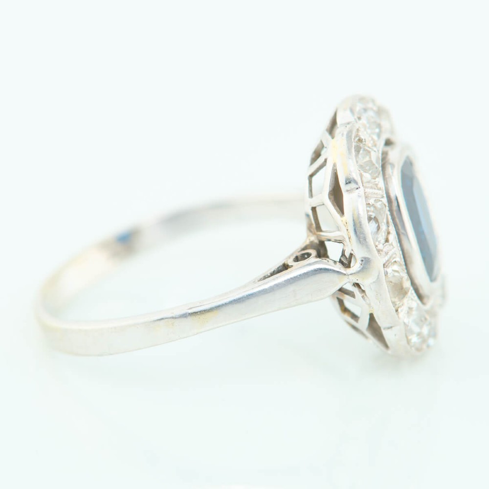 An 18ct white gold cluster ring featuring a central cushion cut blue sapphire framed by a halo of old cut diamonds with millgrain detailing. View IV