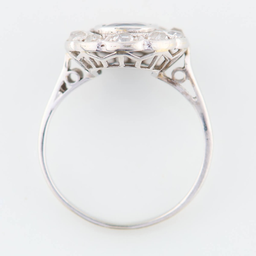 An 18ct white gold cluster ring featuring a central cushion cut blue sapphire framed by a halo of old cut diamonds with millgrain detailing. View V