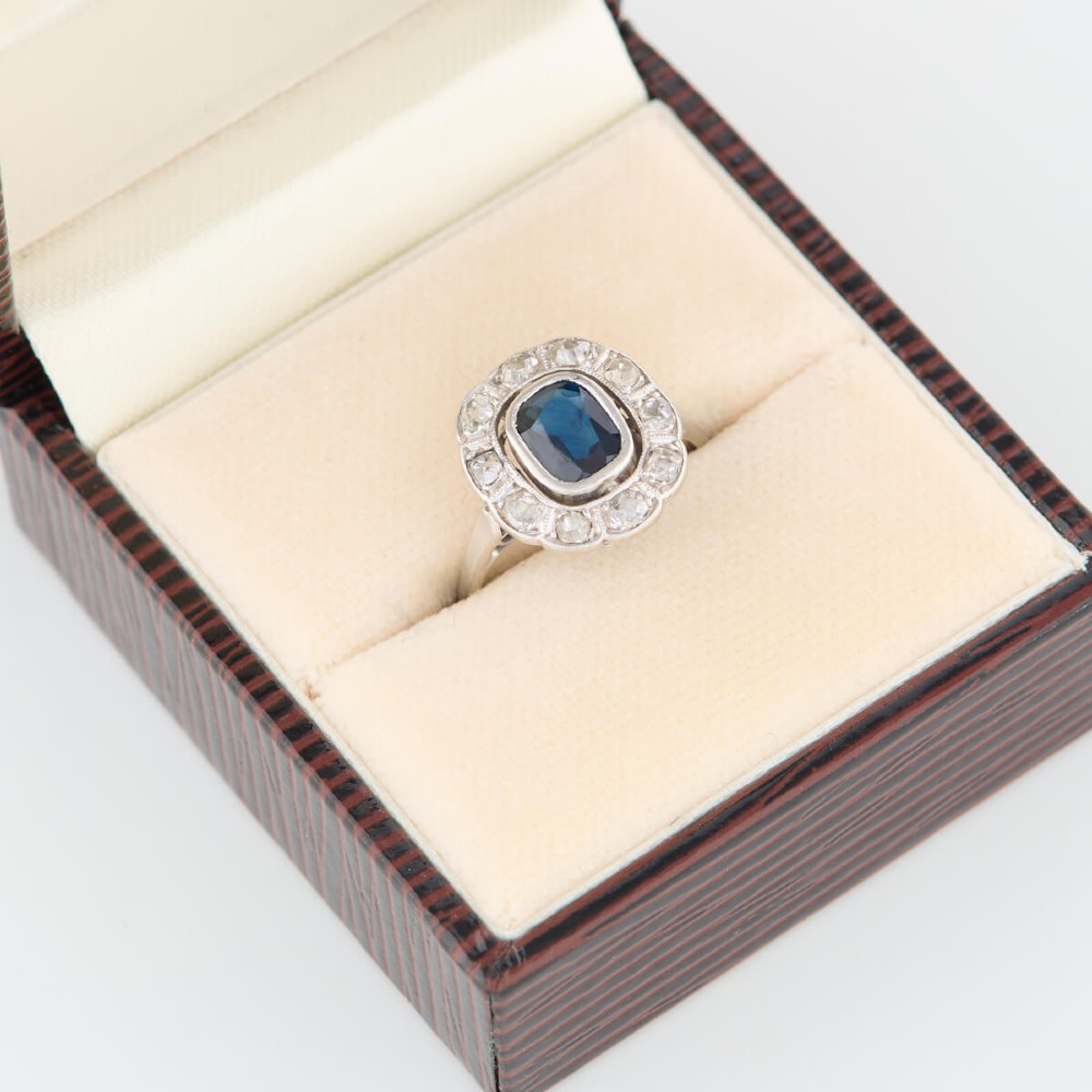 An 18ct white gold cluster ring featuring a central cushion cut blue sapphire framed by a halo of old cut diamonds with millgrain detailing. View VI