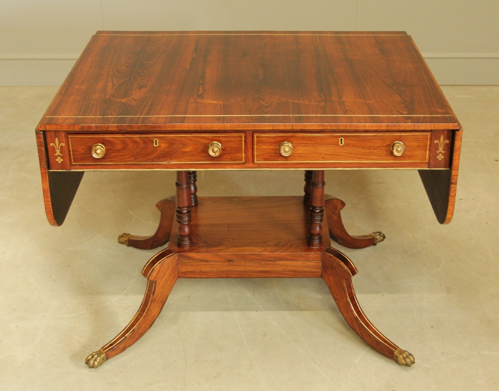 A Regency rosewood sofa table featuring brass stringing inlay, two frieze drawers, twin turned pedestal columns, and splayed legs with brass paw feet.