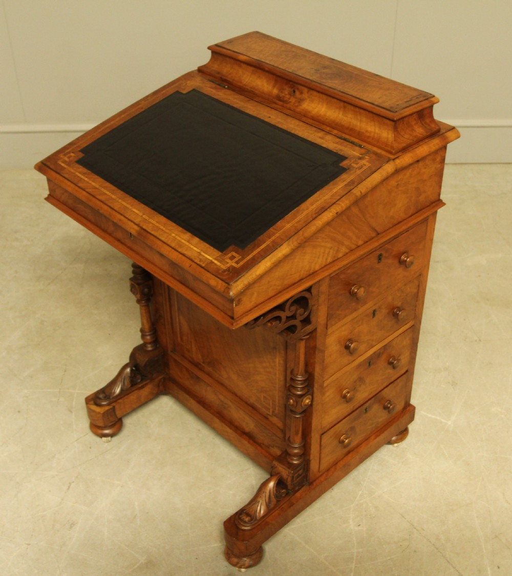 A Victorian walnut Davenport desk featuring an inlaid sloped dark leather writing surface, side drawers with wooden handles, carved front support columns, and brass castors. View II
