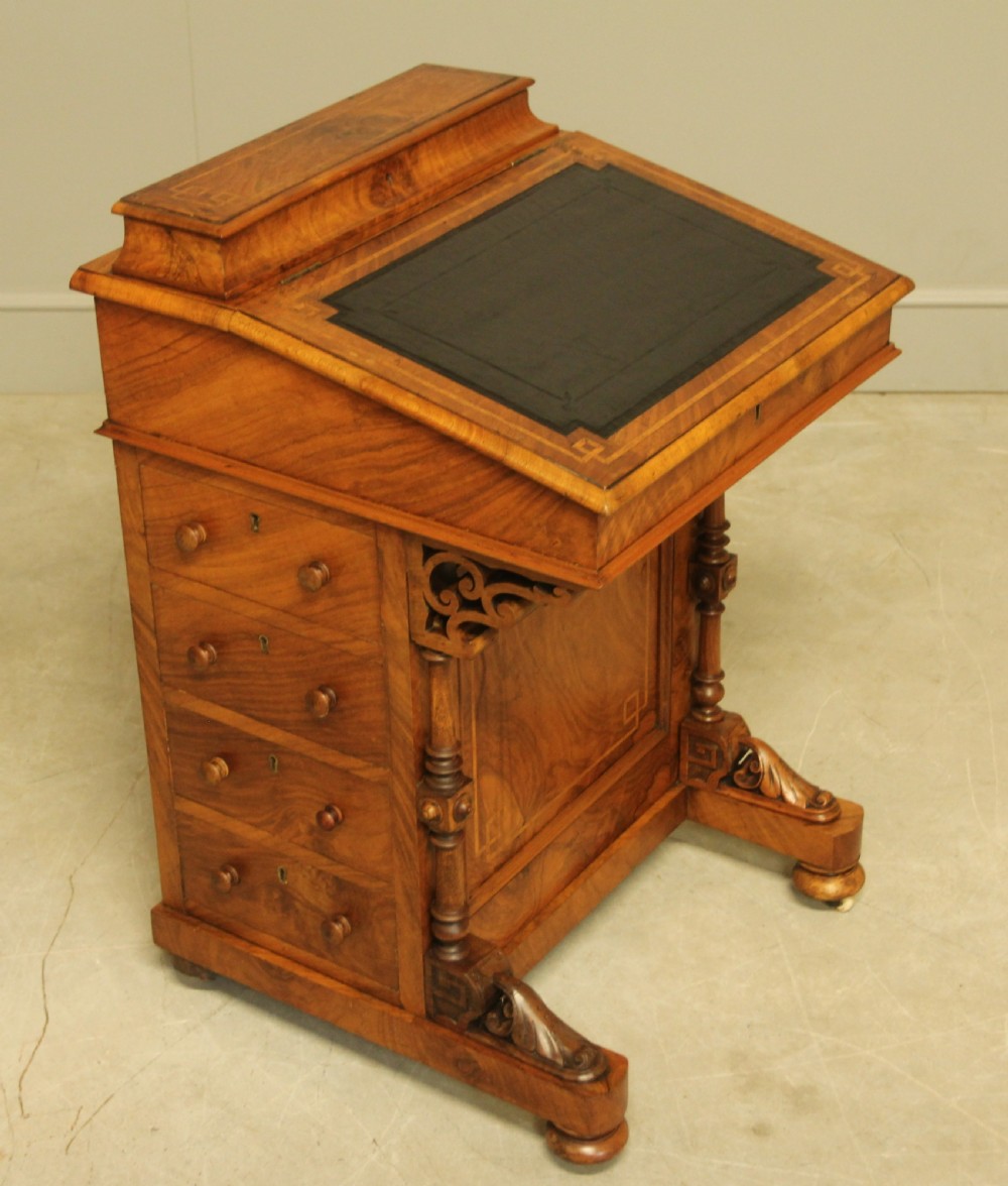 A Victorian walnut Davenport desk featuring an inlaid sloped dark leather writing surface, side drawers with wooden handles, carved front support columns, and brass castors. View VI