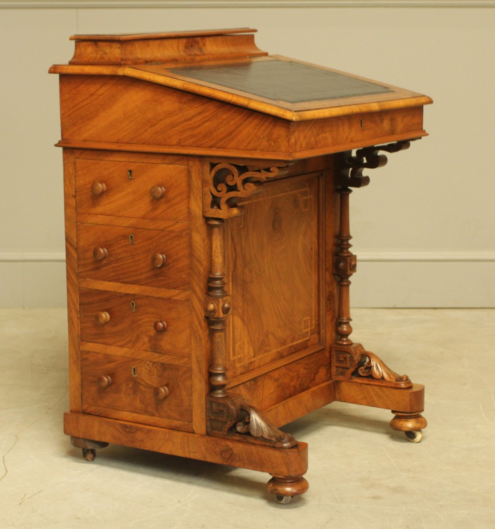 A Victorian walnut Davenport desk featuring an inlaid sloped dark leather writing surface, side drawers with wooden handles, carved front support columns, and brass castors. View VII