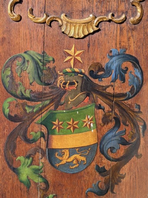 fig 1. A rectangular carved dark oak panel featuring a hand-painted heraldic coat of arms with a Latin motto and gilded rococo scrollwork border.