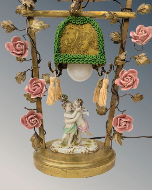 fig 1. An antique French gilt bronze table lamp featuring an arched frame decorated with pink porcelain roses, enclosing a pair of ceramic cherubs and a fabric-trimmed shade.