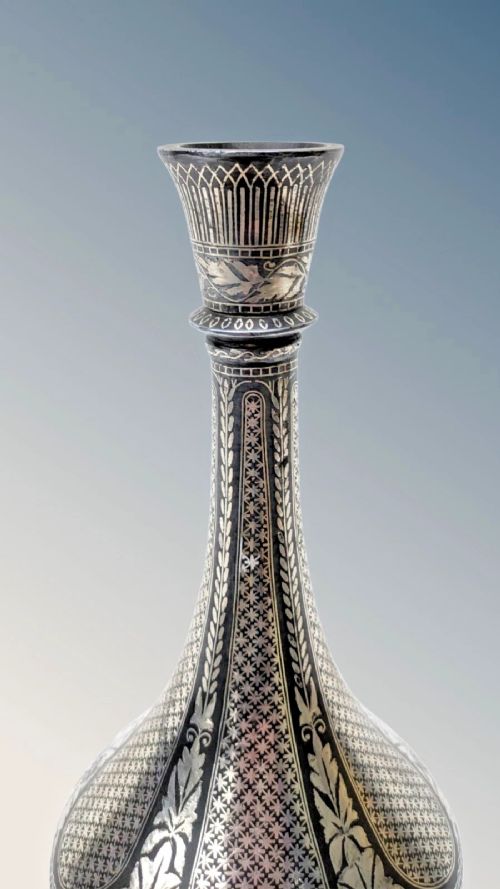 fig 1. An Indian Bidriware metal vase featuring a bulbous body, long neck, flared rim, and intricate silver inlay in floral and geometric patterns against a dark black ground.