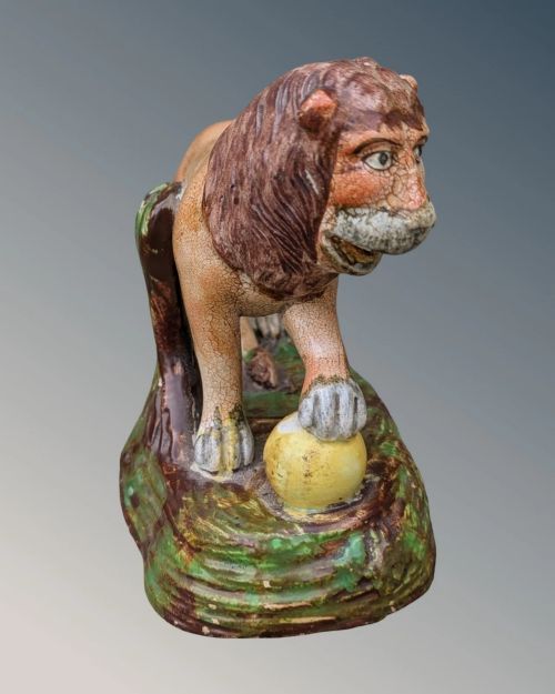 fig 1. An early nineteenth-century Staffordshire pearlware lion figure featuring an ochre-tinted body with one paw resting on a yellow sphere, set upon a moulded green and brown glaze base.