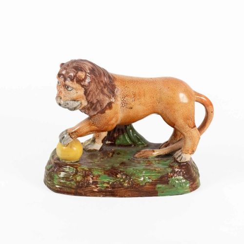 An early nineteenth-century Staffordshire pearlware lion figure featuring an ochre-tinted body with one paw resting on a yellow sphere, set upon a moulded green and brown glaze base. View X