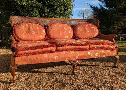 fig 1. An antique wooden three-seater sofa featuring a woven cane backrest, carved legs, and soft terracotta floral cushions with fringe trim.
