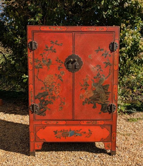 fig 2. A red lacquered two-door Chinese cabinet decorated with black and gold hand-painted floral and bird motifs, supported on straight block feet and fitted with brass hardware.