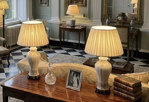 fig 3. A pair of white porcelain table lamps decorated with ornate gold leaf detailing, mounted on brass bases, and topped with cream pleated fabric lampshades.
