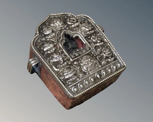 fig 2. A Tibetan silver Gau travelling prayer box featuring embossed repousse ornamentation, an arched central niche housing a deity, and a copper casing.
