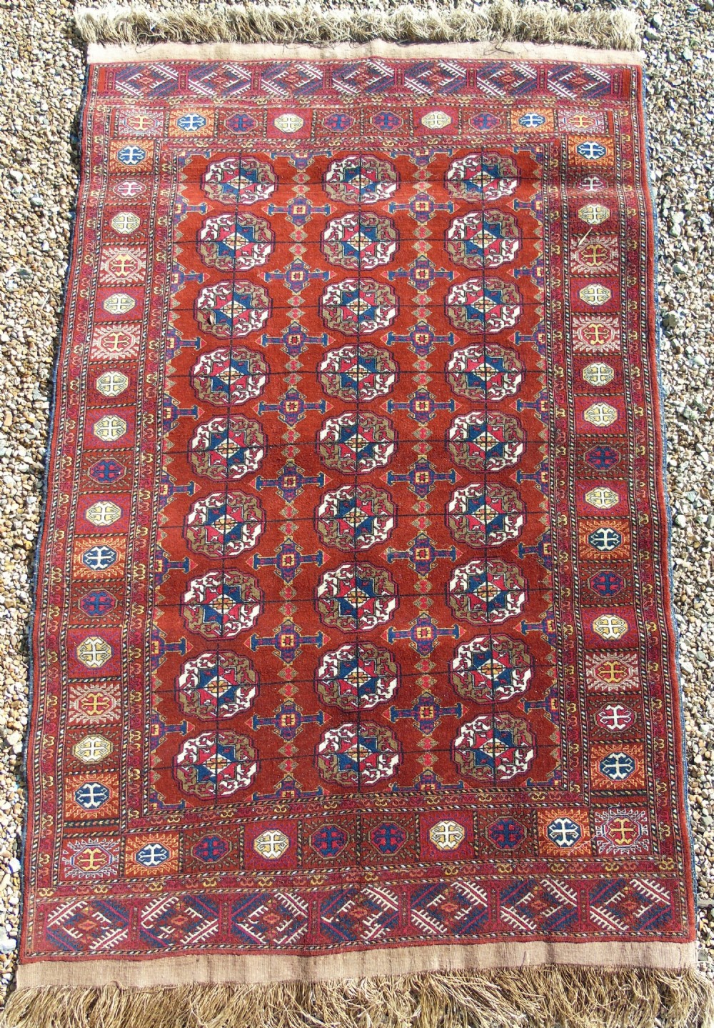 An antique Tekke Turkmen wool carpet featuring repeating octagonal gul motifs across a terracotta red field framed by multiple geometric borders and fringe.