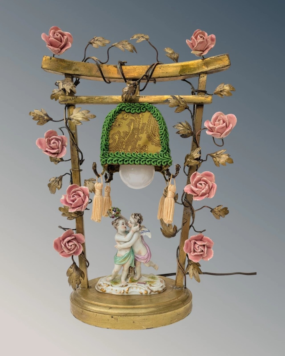 An antique French gilt bronze table lamp featuring an arched frame decorated with pink porcelain roses, enclosing a pair of ceramic cherubs and a fabric-trimmed shade.