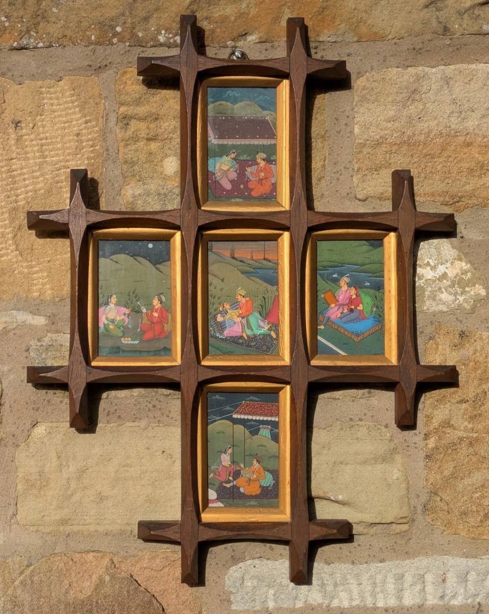 A cruciform dark wooden frame containing five rectangular Mughal style gouache paintings depicting courtly figures in landscape settings.