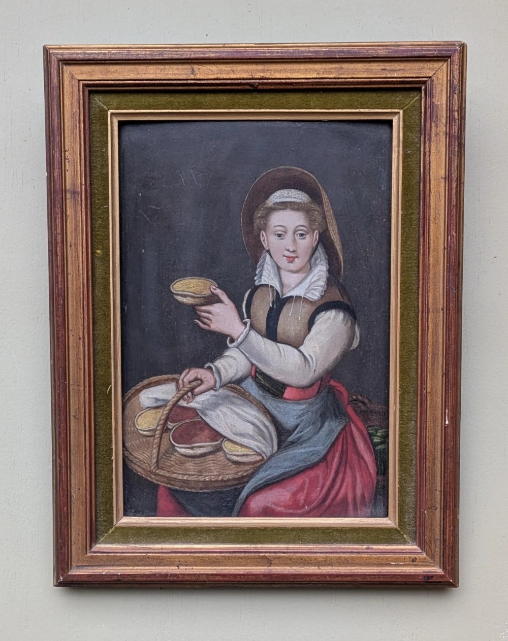 A 19th-century oil painting depicting a woman holding a small bowl, positioned behind a woven basket containing various spices, set within a moulded wooden frame with a velvet slip.