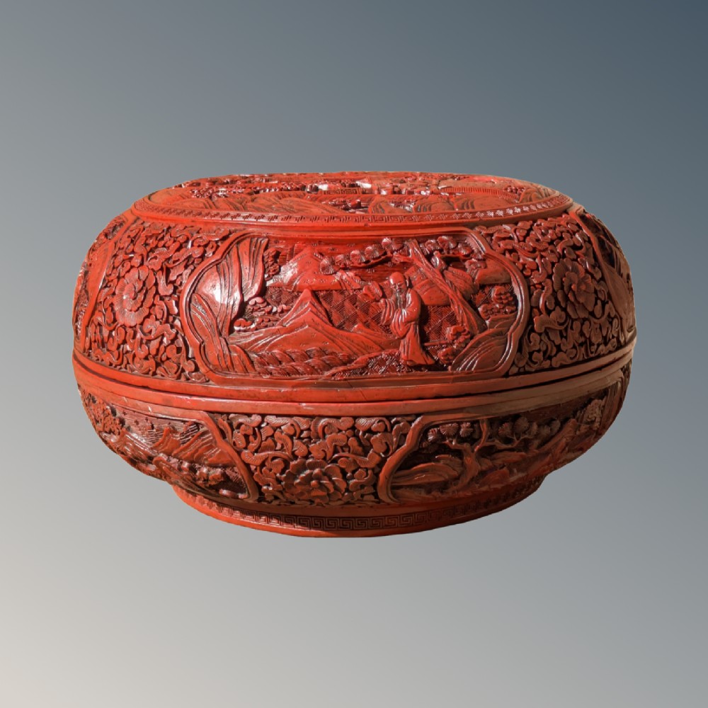 A circular Chinese cinnabar lacquer box features deeply carved relief depictions of figures in mountain landscapes framed by floral and geometric borders.