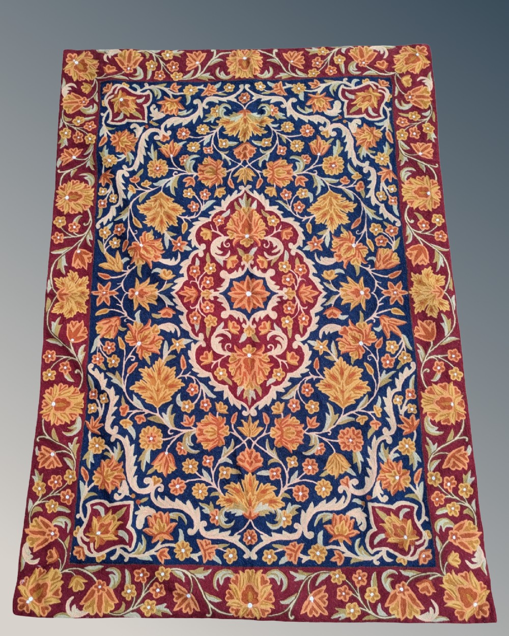 A rectangular Kashmir chain stitch rug featuring dense floral embroidery, a central medallion, and borders in red, navy blue, and gold tones.