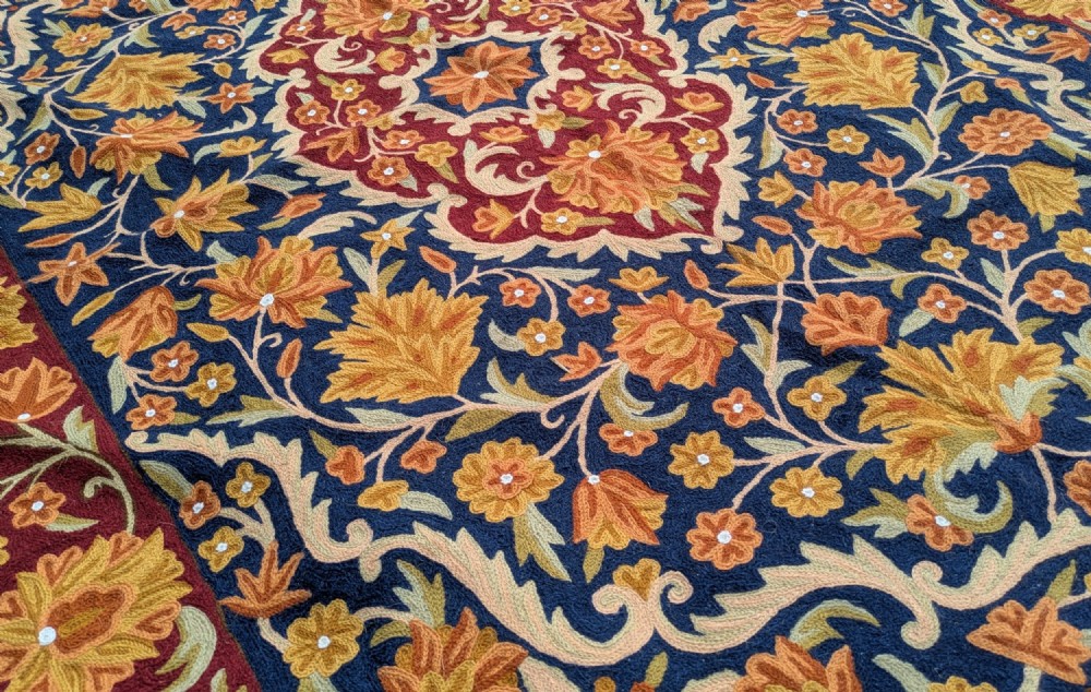 A rectangular Kashmir chain stitch rug featuring dense floral embroidery, a central medallion, and borders in red, navy blue, and gold tones. View IV
