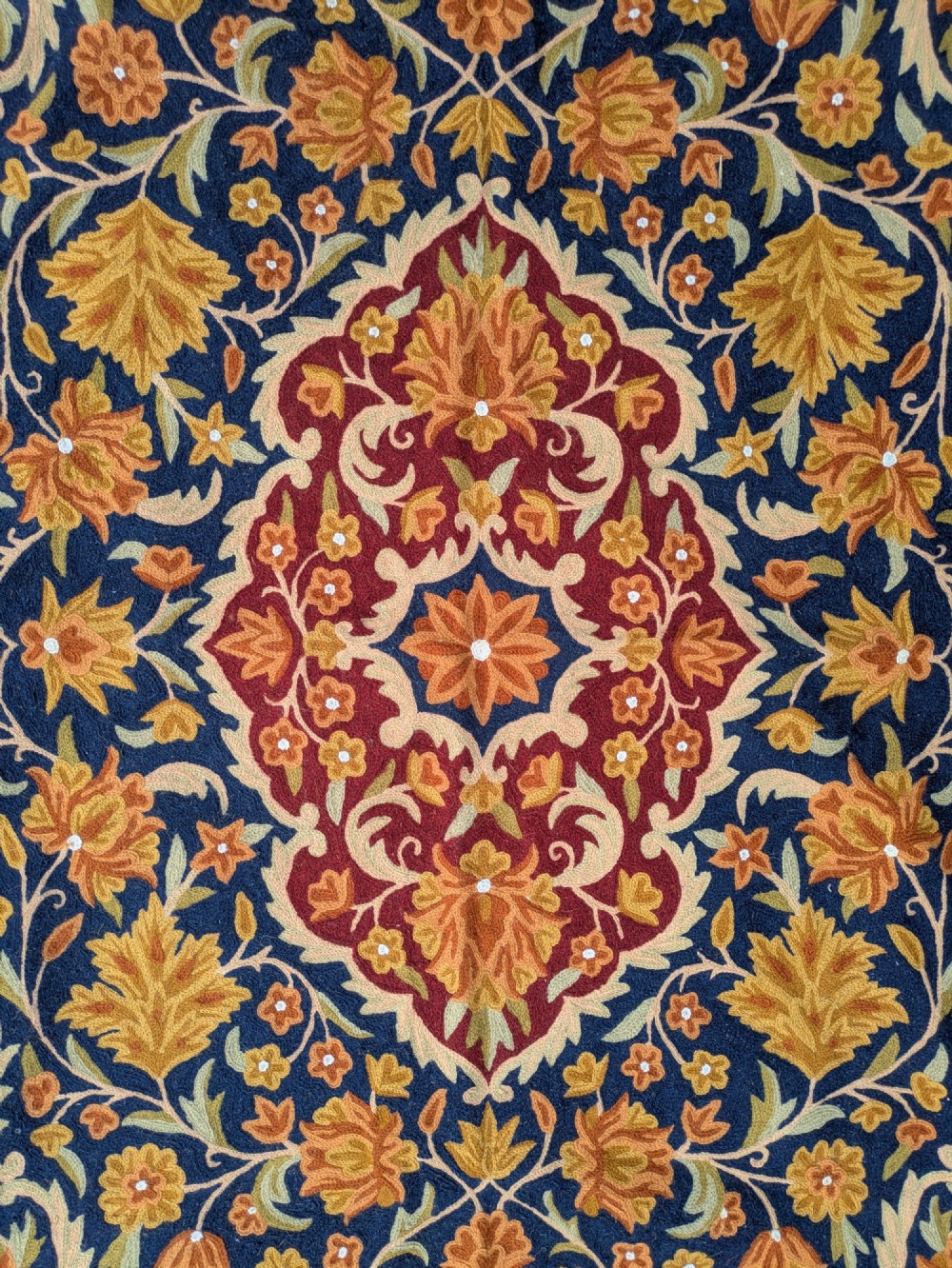 A rectangular Kashmir chain stitch rug featuring dense floral embroidery, a central medallion, and borders in red, navy blue, and gold tones. View V