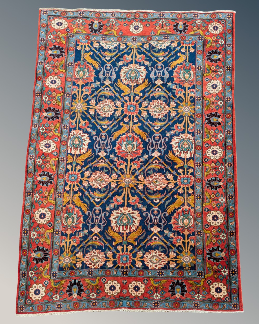 An antique hand-knotted Veramin Persian carpet featuring a dark blue central field filled with a floral lattice pattern and birds, bordered by a wide red band with rosettes.