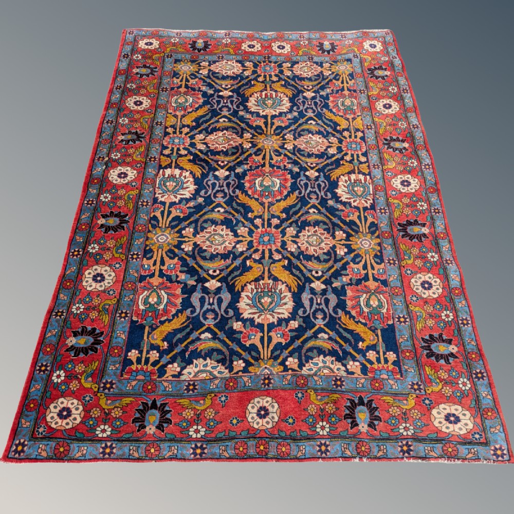 An antique hand-knotted Veramin Persian carpet featuring a dark blue central field filled with a floral lattice pattern and birds, bordered by a wide red band with rosettes. View IV