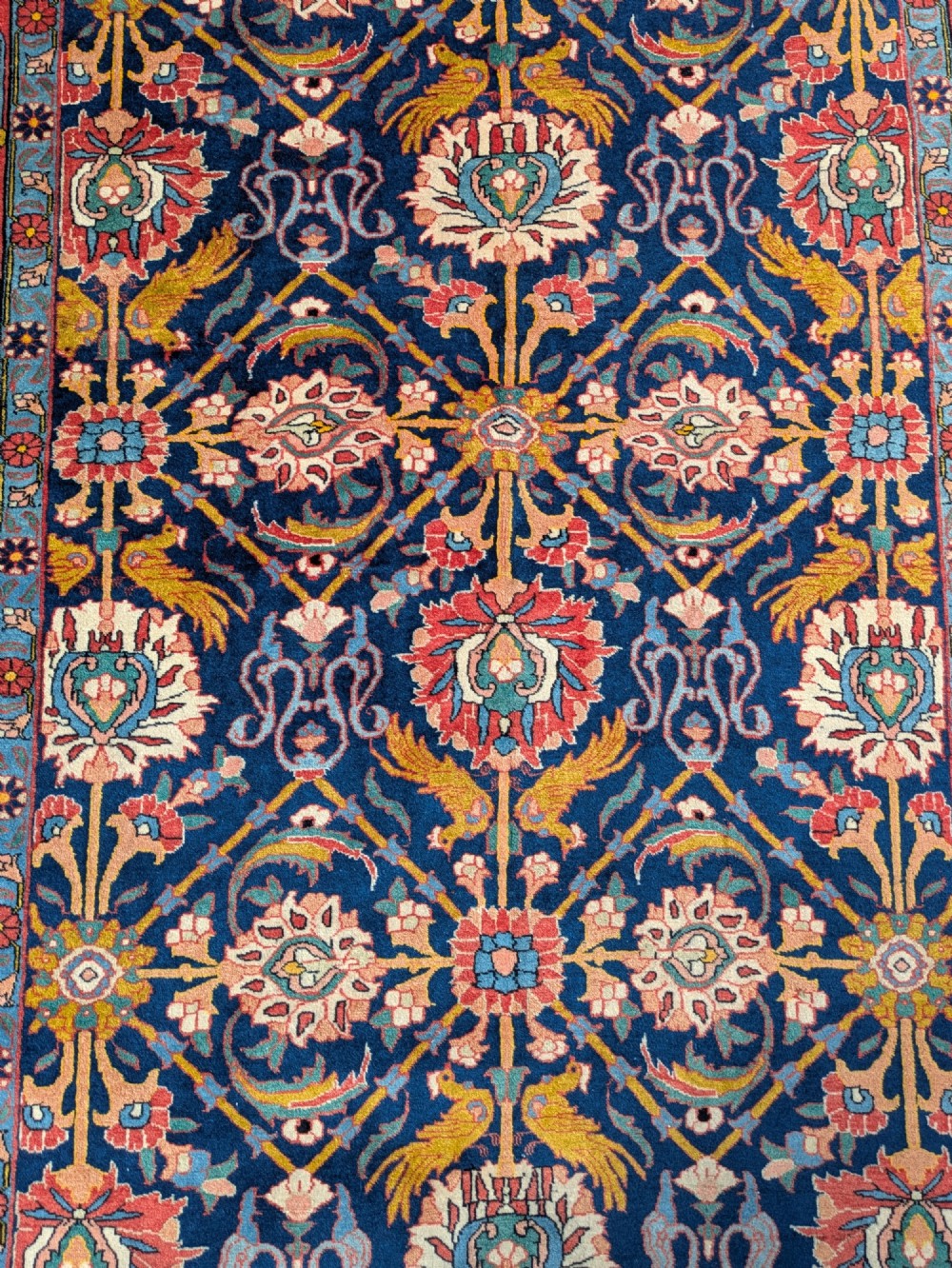 An antique hand-knotted Veramin Persian carpet featuring a dark blue central field filled with a floral lattice pattern and birds, bordered by a wide red band with rosettes. View V