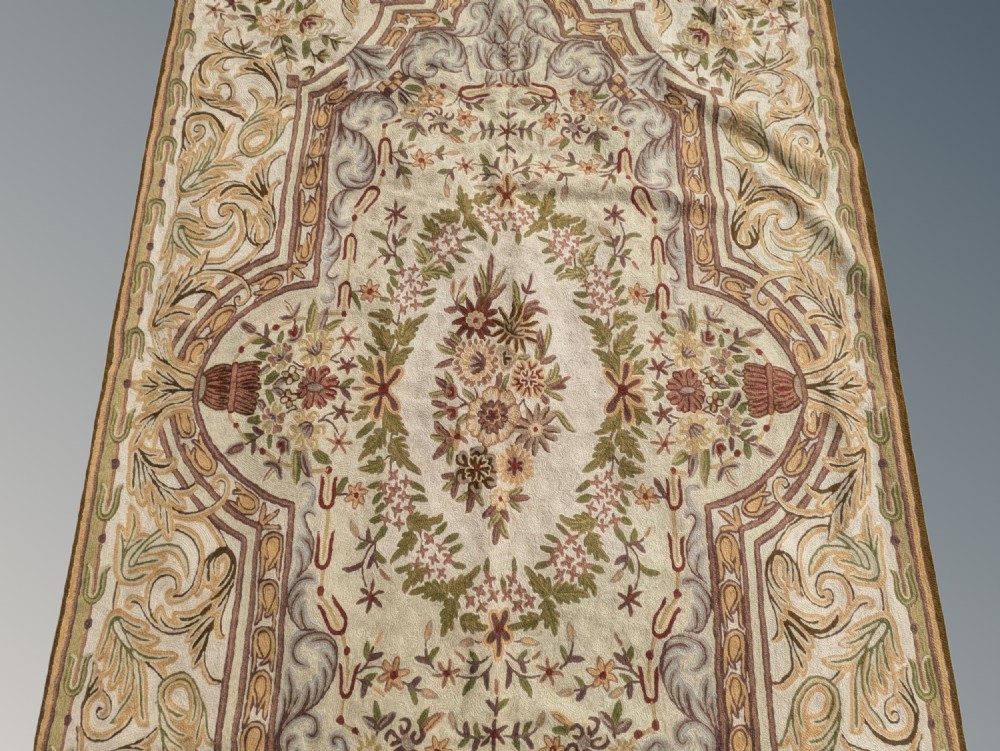 A cream hand-embroidered wool chain-stitch rug featuring a central floral medallion surrounded by scrollwork borders and fringed edges. View II