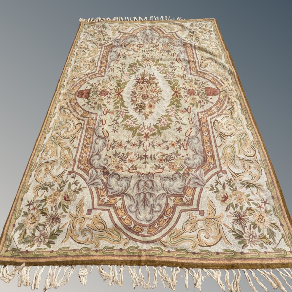 A cream hand-embroidered wool chain-stitch rug featuring a central floral medallion surrounded by scrollwork borders and fringed edges.