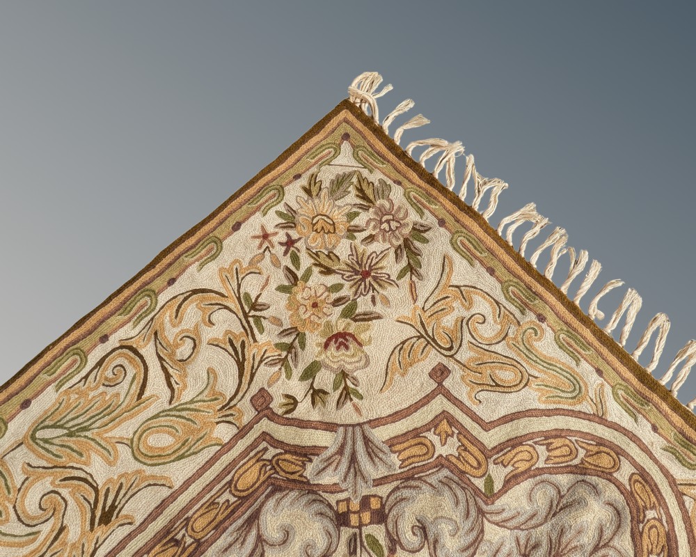 A cream hand-embroidered wool chain-stitch rug featuring a central floral medallion surrounded by scrollwork borders and fringed edges. View III