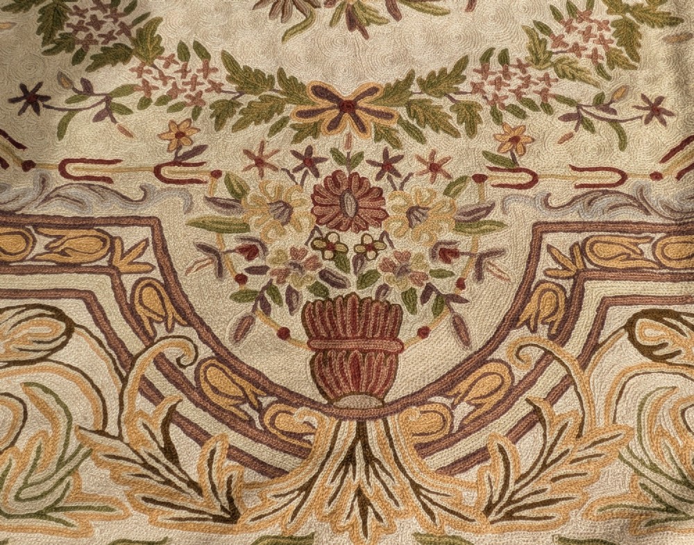 A cream hand-embroidered wool chain-stitch rug featuring a central floral medallion surrounded by scrollwork borders and fringed edges. View IV