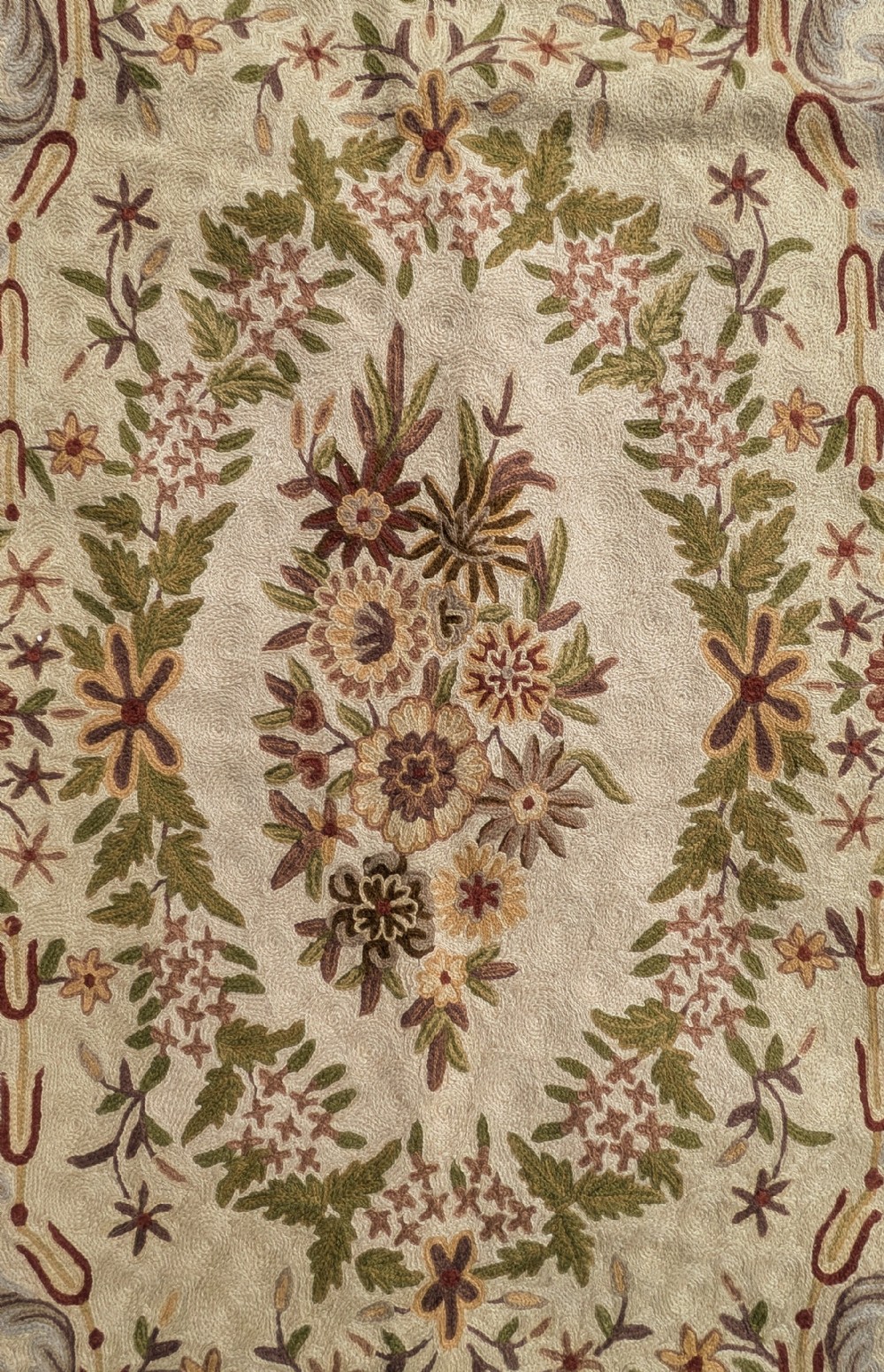 A cream hand-embroidered wool chain-stitch rug featuring a central floral medallion surrounded by scrollwork borders and fringed edges. View V