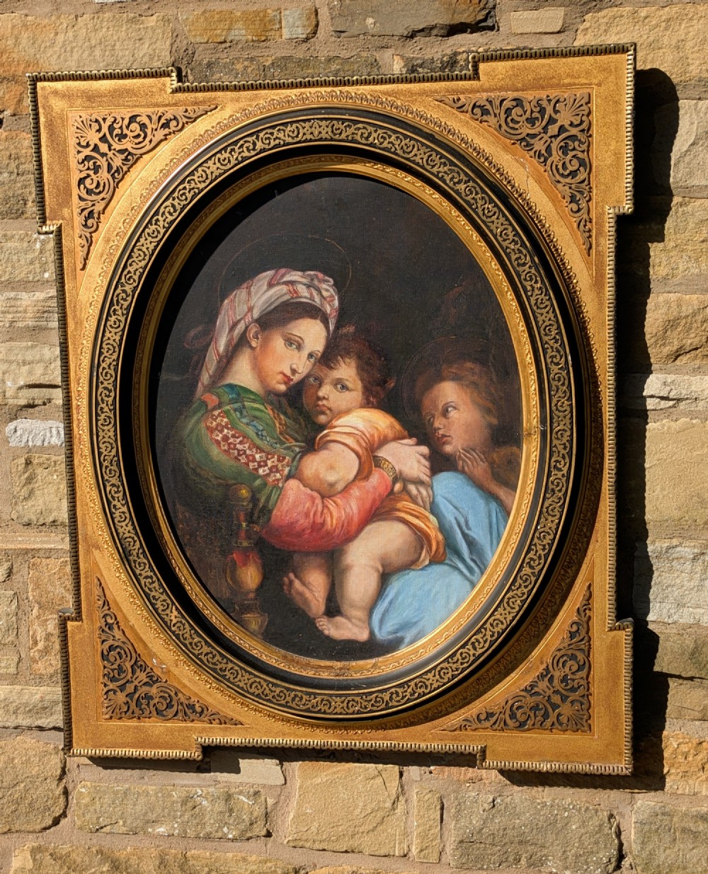 An oval oil painting depicting Mary holding the infant Christ alongside Saint John, set within an ornate gilded frame with corner fretwork.
