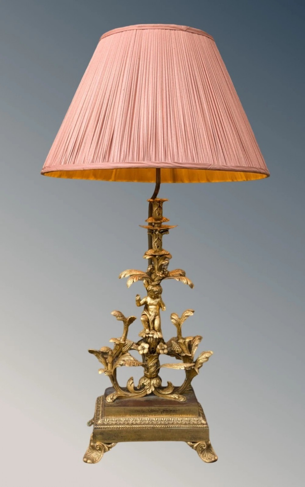 An ornate gilt bronze table lamp featuring a sculpted cherub figure on a floral cast base with a pleated pink silk shade.