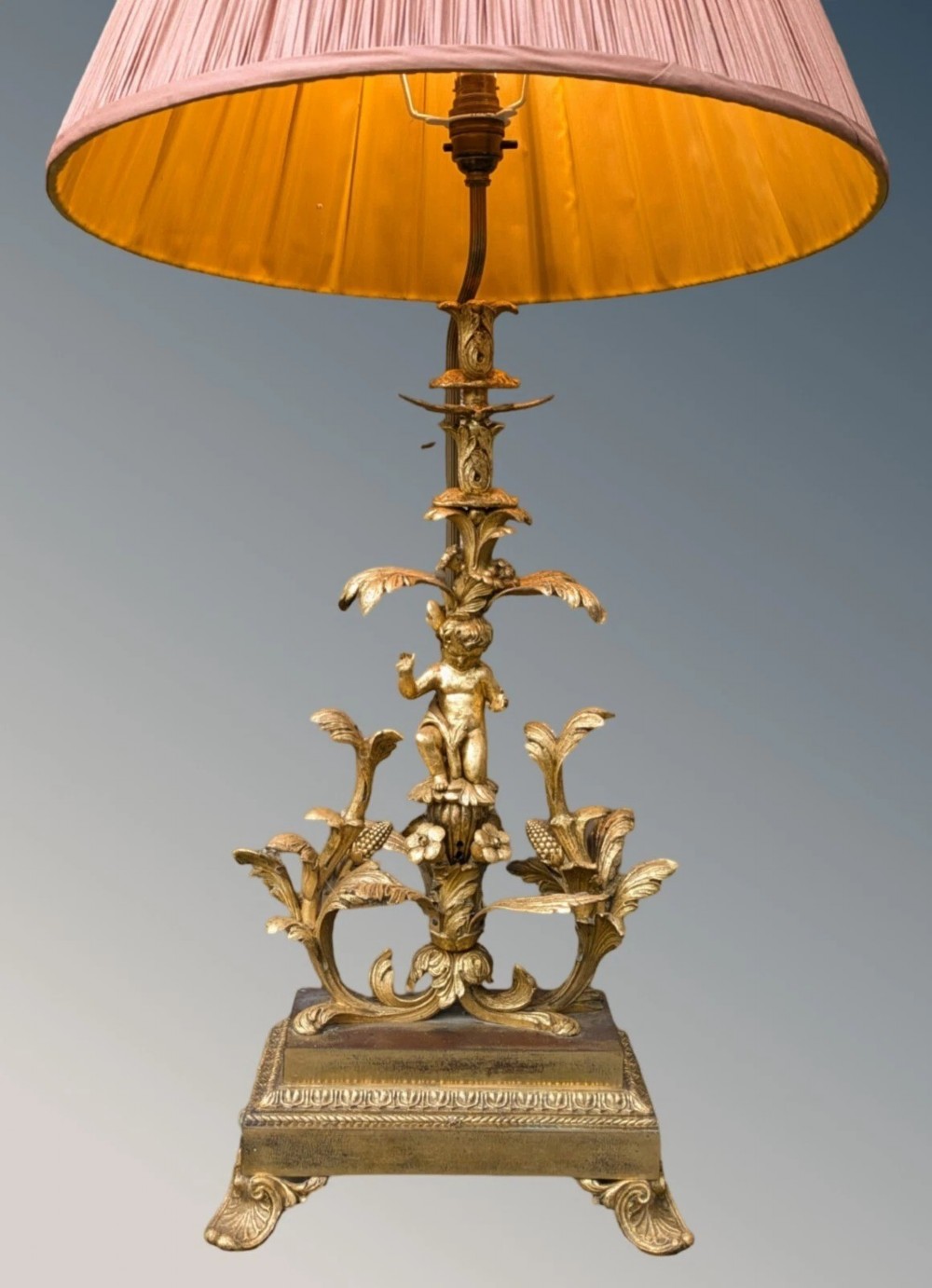 An ornate gilt bronze table lamp featuring a sculpted cherub figure on a floral cast base with a pleated pink silk shade. View II