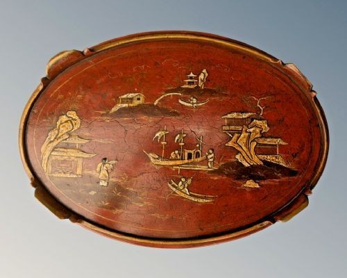An oval antique red lacquer side table featuring hand-painted gold chinoiserie narrative scenes on the surface and slender legs. View IV