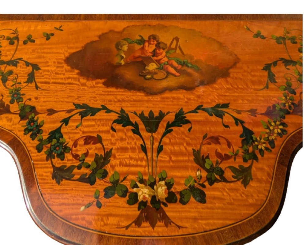 A Regency satinwood side table with a shaped top decorated with hand-painted floral swags and a central cherub medallion, supported by slim tapering legs. View V
