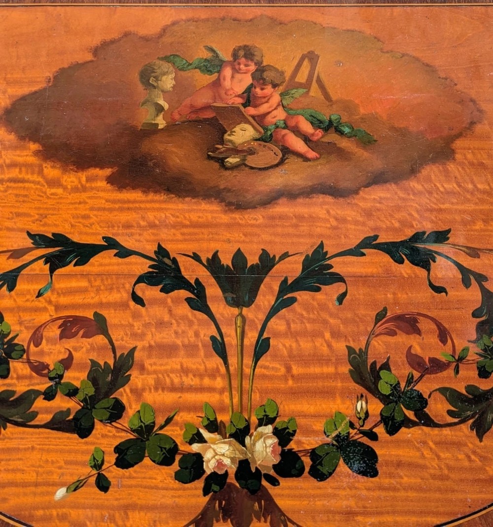 A Regency satinwood side table with a shaped top decorated with hand-painted floral swags and a central cherub medallion, supported by slim tapering legs. View IX