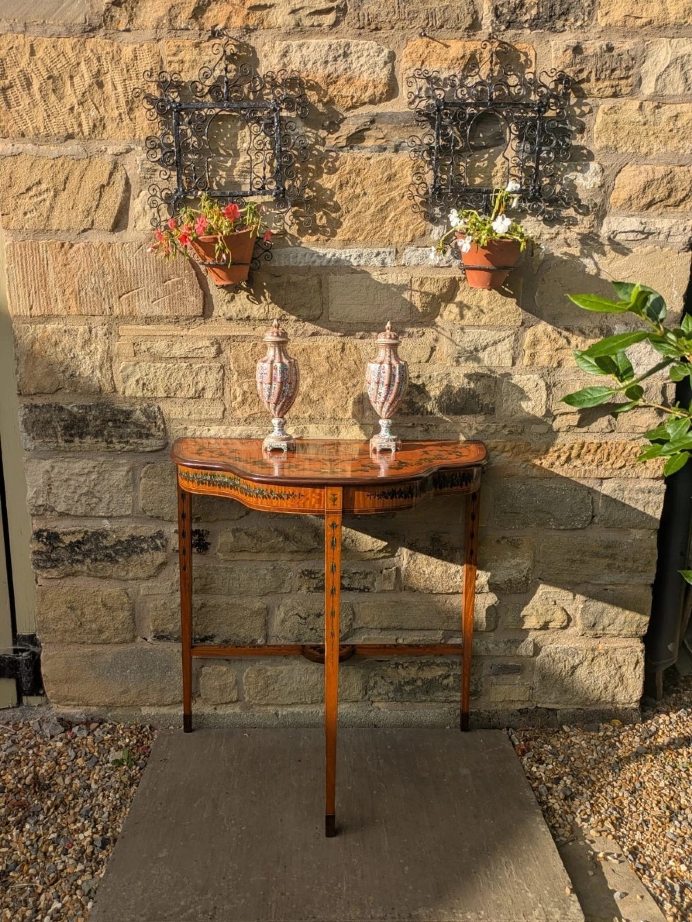 A Regency satinwood side table with a shaped top decorated with hand-painted floral swags and a central cherub medallion, supported by slim tapering legs. View X