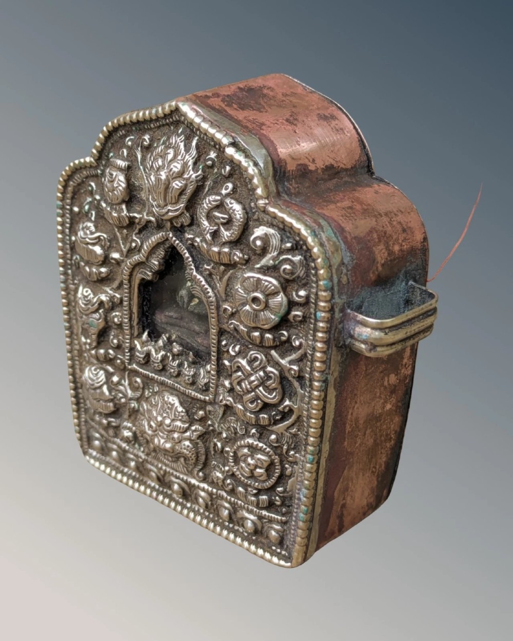 An arched Tibetan Gau prayer shrine featuring an intricately embossed silver metal front panel mounted on a copper casing with side loops.
