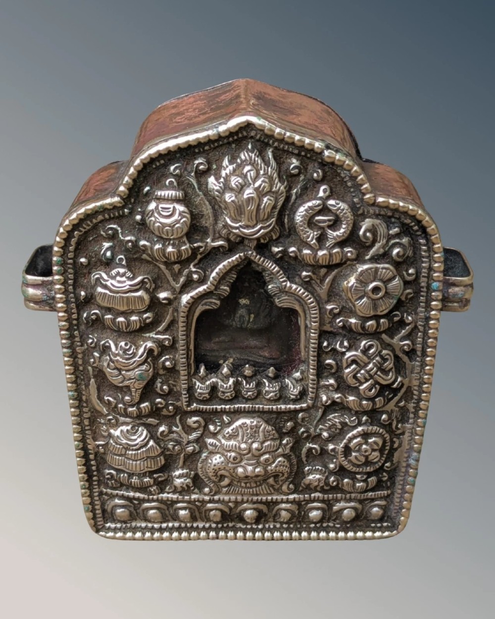 An arched Tibetan Gau prayer shrine featuring an intricately embossed silver metal front panel mounted on a copper casing with side loops. View II